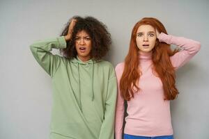 Indoor photo of young pretty bemused women clutching their heads with raised palms while looking at camera with puzzled faces, posing over grey background in comfortable wear