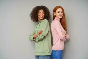 Studio photo of cheerful pretty young ladies posing back to back over grey background and keeping their hands folded, being in nice mood and smiling positively