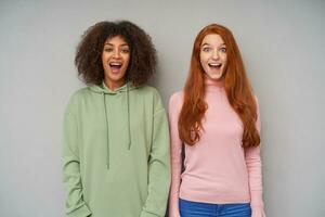 Studio shot of two beautiful joyful young ladies looking excitedly at camera and keeping their mouths wide opened, standing over grey background with hands down photo