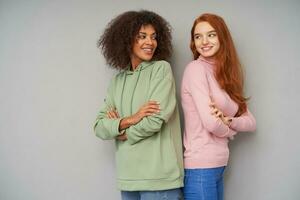 Positive young beautiful women looking cheerfully over thier shoulders on each other and smiling gladly, standing over grey background with crossed hands photo