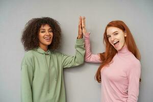 Cheerful young attractive ladies raising their palms and giving five to each other while looking positively at camera with wide smiles, isolated over grey background photo
