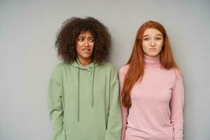 Puzzled young pretty ladies dressed in comfortable wear grimacing their faces and frowning confusedly eyebrows while standing over grey background with hands down photo