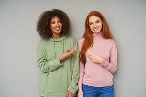 Charming young cheerful ladies showing their white perfect teeth while smiling positively and pointing on each other with raised forefingers, posing over grey background photo