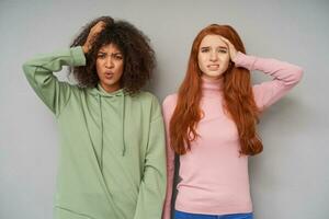 Puzzled young beautiful girlfriends grimacing their faces while looking confusedly at camera, keeping raised hands on their heads while posing over grey background photo