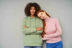 Horizontal photo of upset young pretty women crossing their hands on their chests and keeping lips folded while posing over grey background, dressed in casual clothes