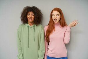 Portrait of young displeased curly dark skinned brunette female in green hoodie frowning her face while posing over grey background with puzzled redhead pretty woman photo