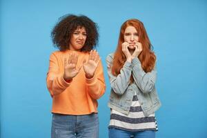 Studio photo of scared curly dark skinned brunette lady raising hands in protection gesture while looking at camera with frightened face, posing over blue background with afraid redhead friend