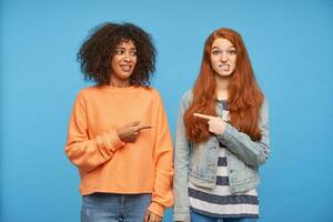 Confused young attractive ladies showing on each other with raised forefingers and frowning eyebrows with embarrassed faces while standing over blue background photo