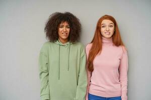 Confused young beautiful women keeping their hands along bodies while standing over grey background, biting underlips and looking at camera with puzzled faces photo