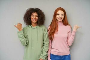 Glad attractive young ladies dressed in casual wear showing with their thumbs in different directions and looking cheerfully at camera with positive smile, isolated over grey background photo