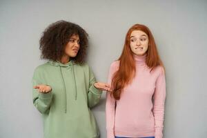 Bewildered young attractive brown haired curly dark skinned woman frowning her face and raising confusedly hands while posing over grey background with puzzled redhead pretty female photo