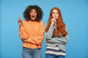 Excited young pretty brown haired dark skinned woman raising forefinger with ideas gesture while posing over blue background with her puzzled redhead friend photo