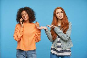 Bewildered young lovely redhead woman showing confusedly with raised hands to her positive brown haired dark skinned brunette friend with mobile phone, isolated over blue background photo