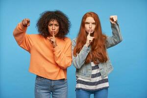 Studio shot of serious young attractive girlfriends showing at camera with raised index fingers while looking severely at camera, asking to be quiet while posing over blue background photo