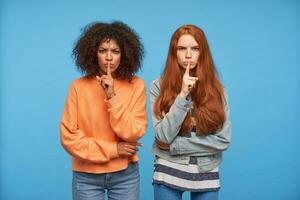 Severe young pretty girlfriends frowning their eyebrows while looking seriously at camera, keeping index fingers on their lips while asking to keep silence, isolated over blue background photo