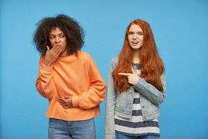 Indoor photo of young brown haired dark skinned female in orange knitted sweater and jeans covering her mouth and holding hand on her belly, posing over blue background with confused redhead lady