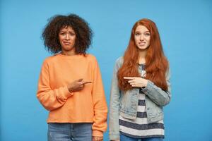 Puzzled young pretty women pointing to each other with index fingers and frimacing confusedly faces while looking at camera, isolated over blue background photo