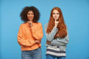 Indoor shot of lovely positive ladies raising hands with hush gesture to their mouths and smiling pleasantly, standing over blue background in casual wear photo