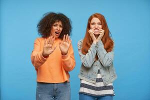Afraid young beautiful ladies grimacing scaredly thier faces and frowning eyebrows while raising hands while standing over blue background with raised hands photo