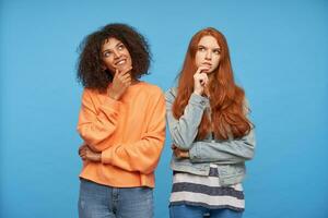Studio photo of thoughtful young pretty ladies looking thoughtfully upwards and holding their chins with raised hands while standing over blue background