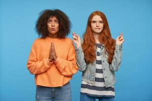 Studio photo of desperate beautiful young women dressed in casual wear raising their hands in hoping gesture and making their wishes, posing over blue background