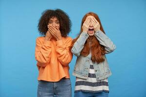 Portrait of brown-eyed curly dark skinned brunette lady covering her mouth with raised hands while standing over blue background with excited joyful redhead female holding palms on her eyes photo