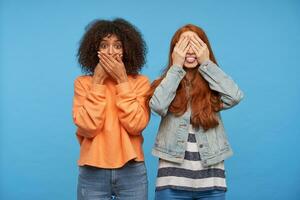Joyful lovely young redhead lady covering her eyes and showing happily tongue while posing over blue background with amazed young dark haired curly dark skinned woman photo