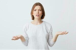 Portrait of confused upset young woman in long-sleeve looks puzzled and holding copy space on both palms isolated over white background photo