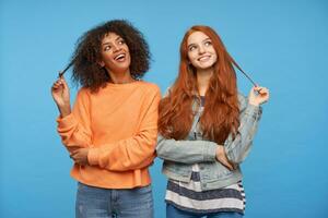 Cheerful beautiful women looking positively upwards and smiling widely, pulling strands of hair with raised hands while standing against blue background photo