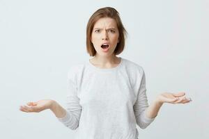 Angry puzzled young woman with opened mouth in long-sleeve looking shocked and holding copy space on both palms isolated over white background photo