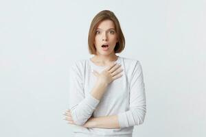 Amazed lovely young woman in long-sleeve standing with opened mouth and hands folded isolated over white background Feels shocked photo