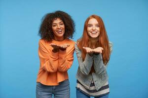 Indoor shot of happy young attractive women looking positively at camera with broad pleasant smile and keeping hands raised, isolated over blue background photo