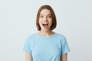 Closeup of amazed excited young woman with opened mouth in blue t shirt looks happy and shouting isolated over white background Feels excited photo