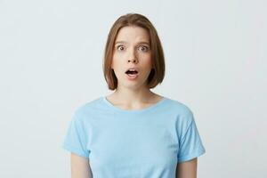 Portrait of amazed beautiful young woman in blue t shirt standing with opened mouth and feels shocked isolated over white background photo