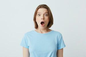 Closeup of shocked stunned young woman in blue t shirt with opened mouth feels amazed and looks directly in camera isolated over white background photo