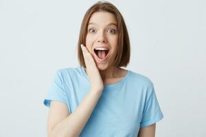 Closeup of happy excited attractive young woman in blue t shirt with opened mouth touching her cheek and feels surprised isolated over white background photo