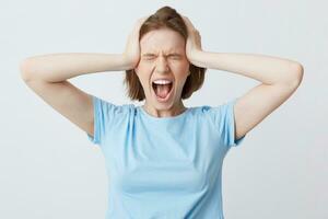 Portrait of crazy hysterical young woman in blue t shirt with closed eyes and hands on head looks stressed and screaming isolated over white background photo