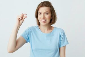 Cute embarrassed pretty young woman in blue t shirt biting her lip and showing tiny size with fingers isolated over white background Looks directly in camera photo