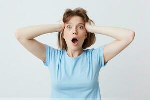 Portrait of astonished crazy young woman in blue t shirt keeps mouth opened and hands on her head isolated over white background Looks shocked and shouting photo