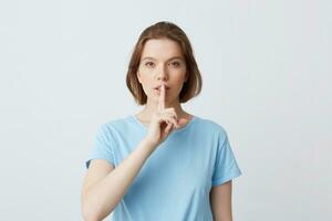 Portrait of peaceful beautiful young woman in blue t shirt looks directly in camera and showing silence gesture isolated over white background photo