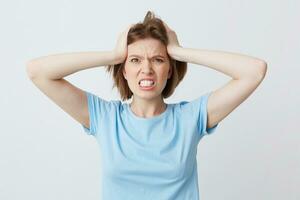 Portrait of angry irritated young woman in blue t shirt standing with hands on head and feels crazy isolated over white background Looks directly in camera photo