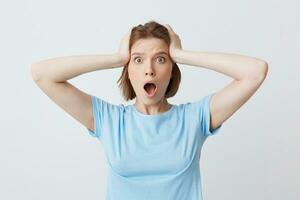 Closeup of amazed shocked young woman in blue t shirt standing with opened mouth and hands on her head isolated over white background Feels stunned and worried photo