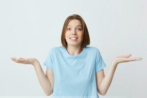 Portrait of confused cute young woman in blue t shirt shrug her shoulders and hold copy space on both palms isolated over white background photo