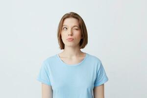 Thoughtful concentrated young woman in blue t shirt with one closed eyes thinking and trying to remember answers for the questions on exam isolated over white wall Looking to the side and up photo