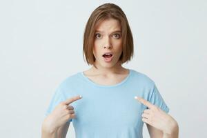 Amazed shocked young woman in blue t shirt with opened mouth looks stunned and points at herself with fingers on both hands isolated over white background photo