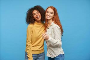Couple of beautiful young ladies dressed in jeans embracing each other and looking happily at camera with charming smiles, standing over blue background photo