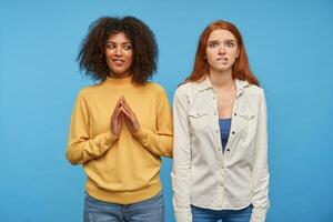 Nervous young pretty redhead female biting her underlip and looking worringly at camera while standing over blue background with her positive lovely dark haired curly dark skinned friend photo