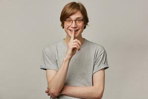 Teenage guy, happy looking man with blond hair. Wearing grey t-shirt, glasses and has braces. People and emotion concept. Showing silence sign. Watching at the camera isolated over grey background photo