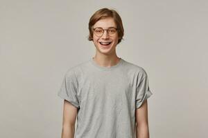 Happy looking man, handsome guy with blond hair. Wearing grey t-shirt, glasses and has braces. People and emotion concept. Watching and broadly smiling at the camera isolated over grey background photo