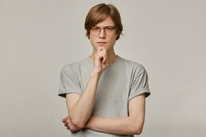 Serious looking male, thinking guy with blond hair. Wearing grey t-shirt and glasses. People and emotion concept. Frowns and touching his chin. Watching at the camera isolated over grey background photo